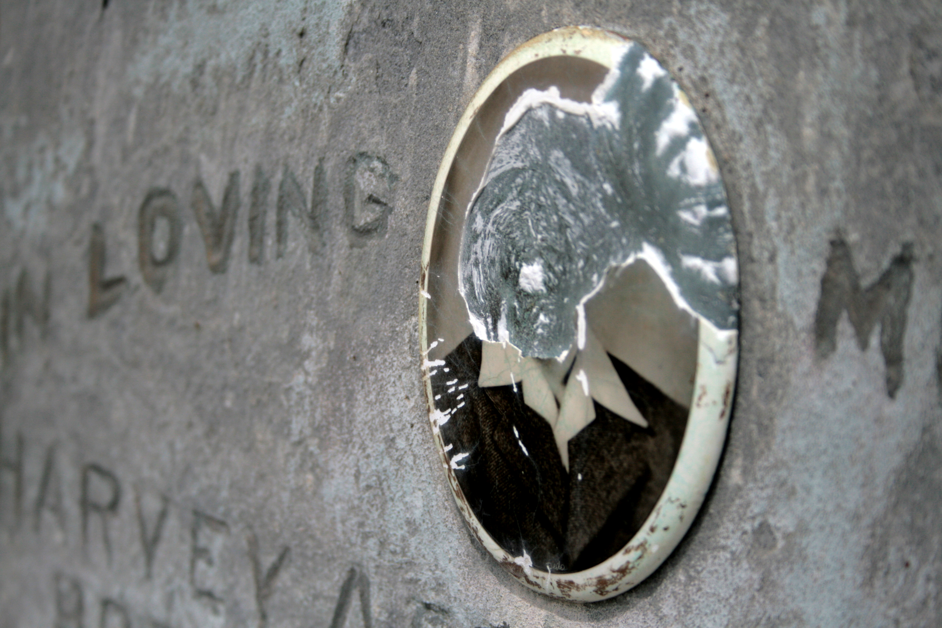 A small oval portrait on a grey tombstone. The portrait is broken and only shows the shoulders of the person in the portriat
